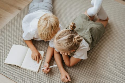 wo children sitting on the floor, working on homework together with notebooks and pencils, surrounded by books and school supplies.