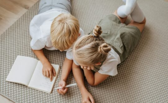 wo children sitting on the floor, working on homework together with notebooks and pencils, surrounded by books and school supplies.