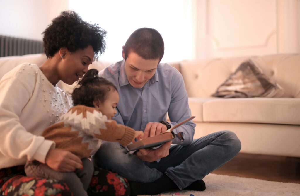 Tow parents male and female reading a book for their little daughter sitting on the floor of the living room next to the couch.