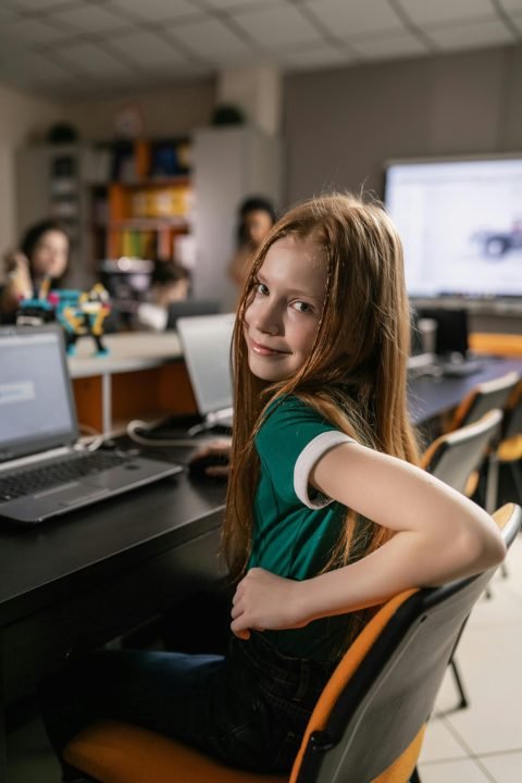 A smiling red-haired girl sits at a classroom desk with a laptop, looking over her shoulder at the camera.