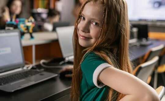 A smiling red-haired girl sits at a classroom desk with a laptop, looking over her shoulder at the camera.