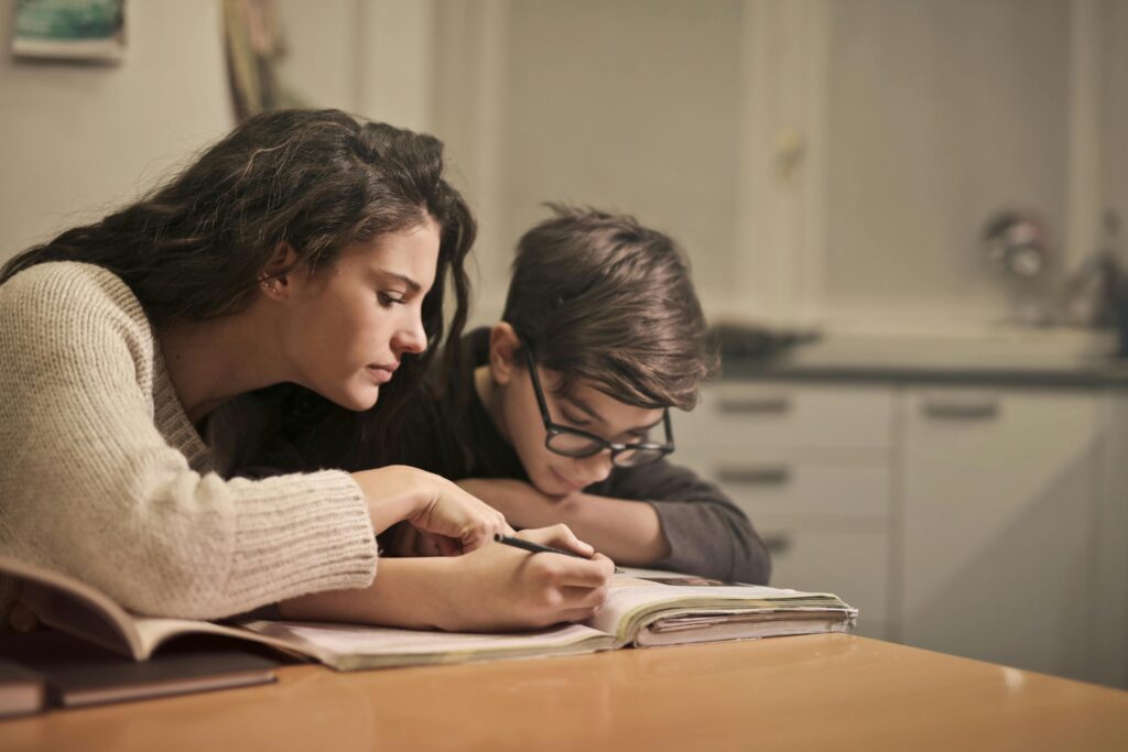 A parent and child sitting at a table, working on homework together with books, notebooks, and a laptop, showcasing a supportive learning moment.