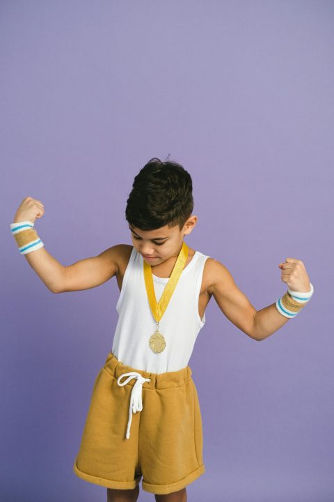 Young boy flexing muscles wearing a medal against a purple background.